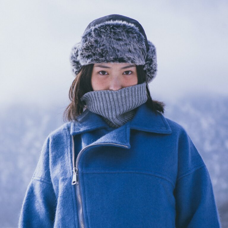 Woman in snowy backdrop with a gray turtleneck, blue leather jacket, and gray ushanka.