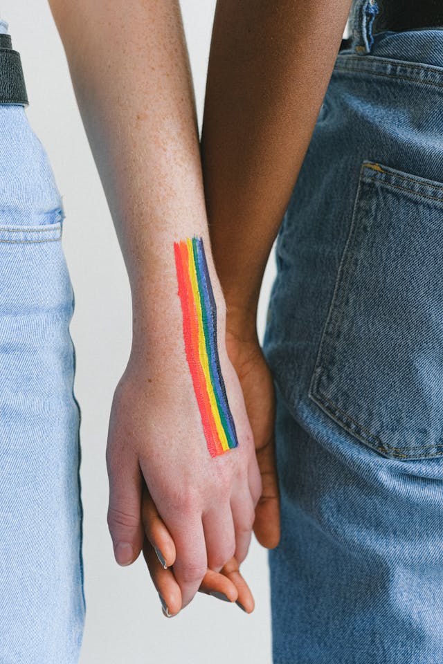 Two LGBTQ+ identifying women in a relationship holding hands with rainbow on left hand.