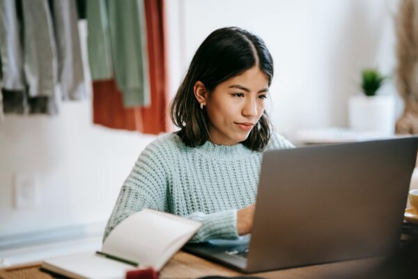 A woman typing on her laptop with an open book next to the laptop.