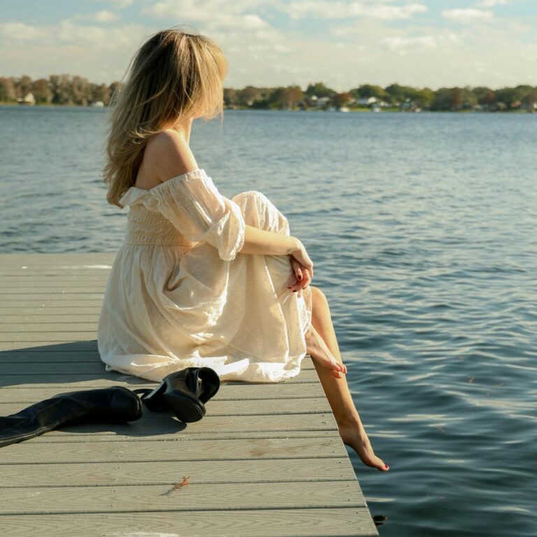 Sideview of a woman with blond hair and white dress is sitting on the edge of a wooden board walk by the sea. Her shoes are placed next to her.