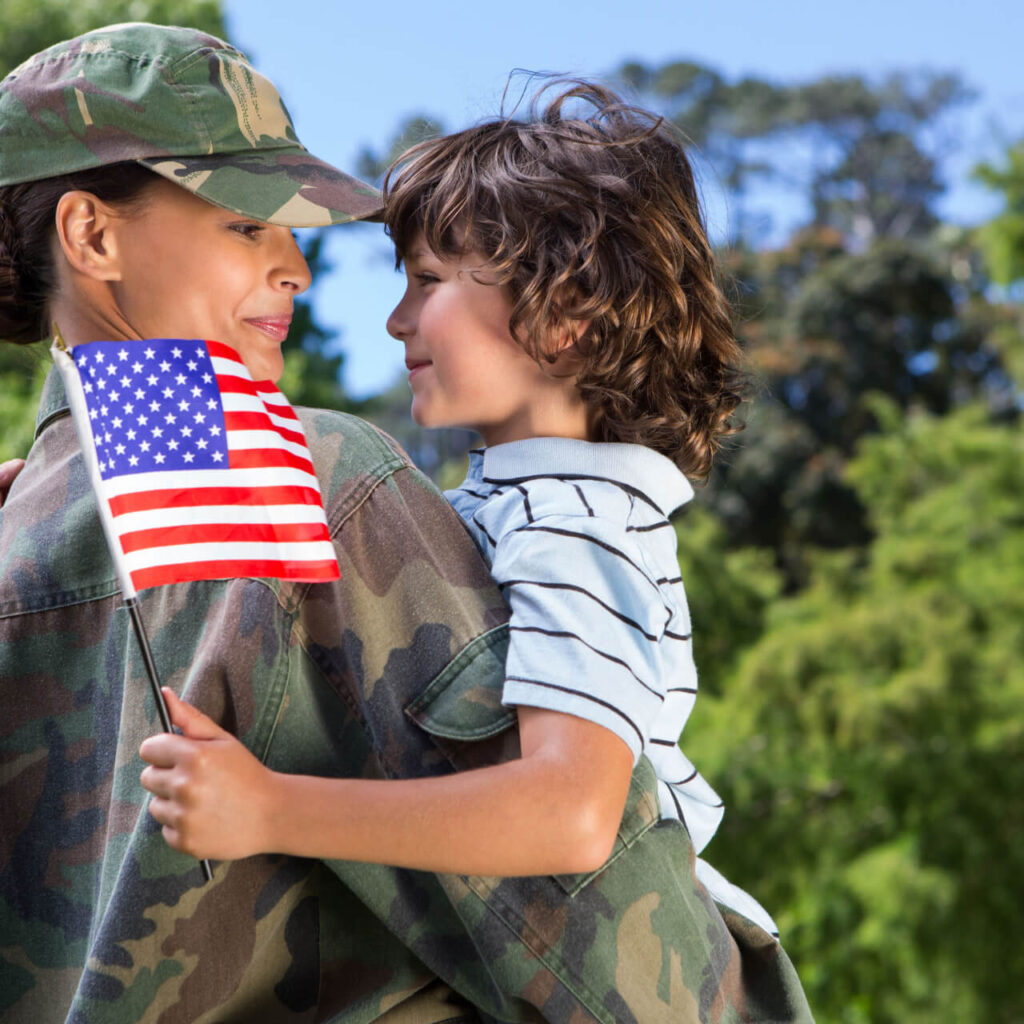 Female veteran in military uniform holding her young child, with the child holding a small American flag.
