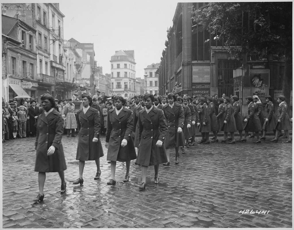 A group of uniformed women from the U.S. Army’s 6888th Central Postal Directory Battalion marches in formation down a cobblestone street in Europe during World War II. Spectators line the sidewalks, watching the parade as the women march in long coats and heeled shoes, displaying discipline and pride. The historic black-and-white photo captures a moment honoring the contributions of the only all-Black, all-female battalion to serve overseas during the war.