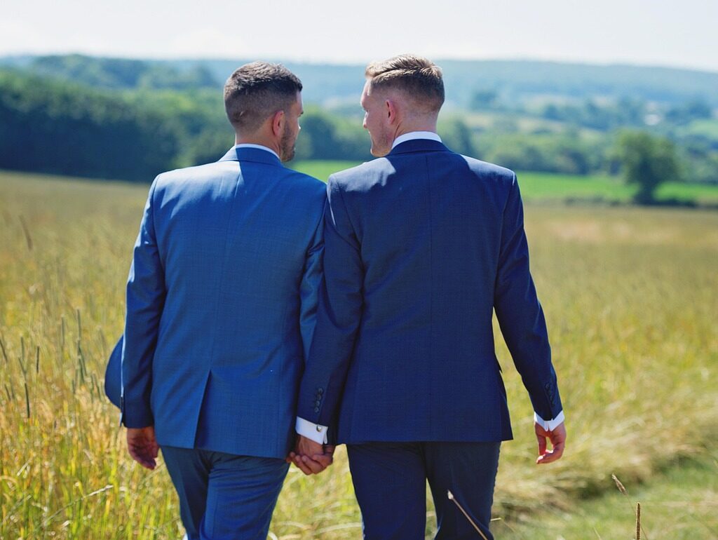 Two men in blue suits walk hand in hand through a grassy field, with green hills and trees in the background.