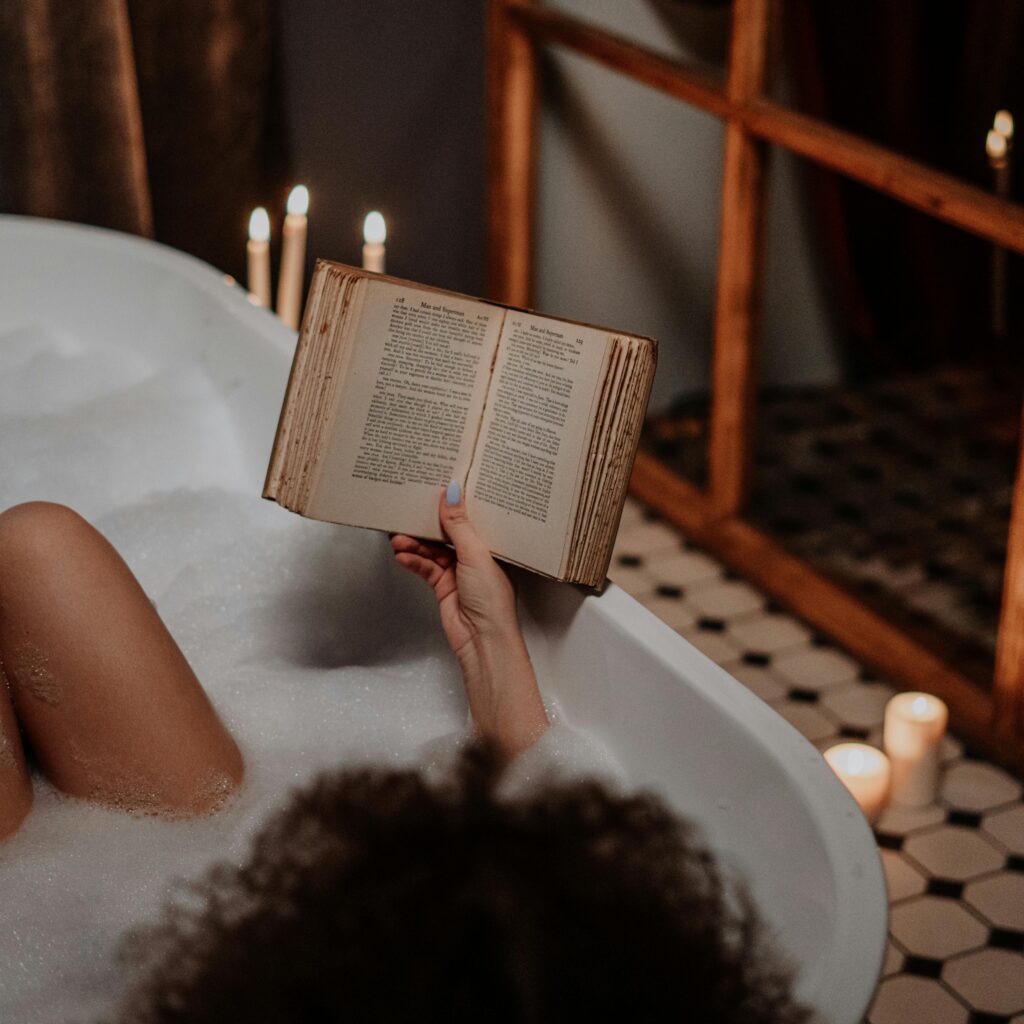 Person relaxing in a bubble bath, holding and reading an old book, with lit candles surrounding the tub for a calm atmosphere.