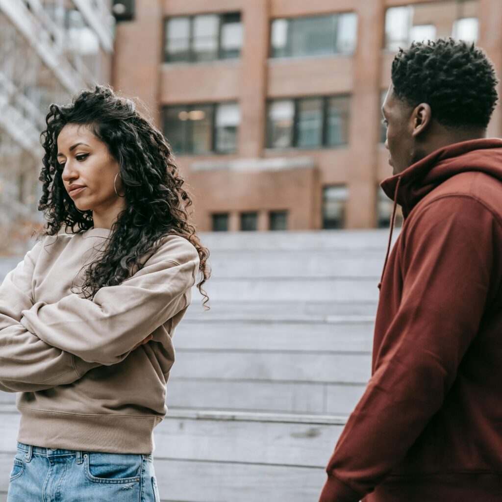Woman with arms crossed looks upset while standing outside, as a man in a red hoodie faces her, suggesting a disagreement.