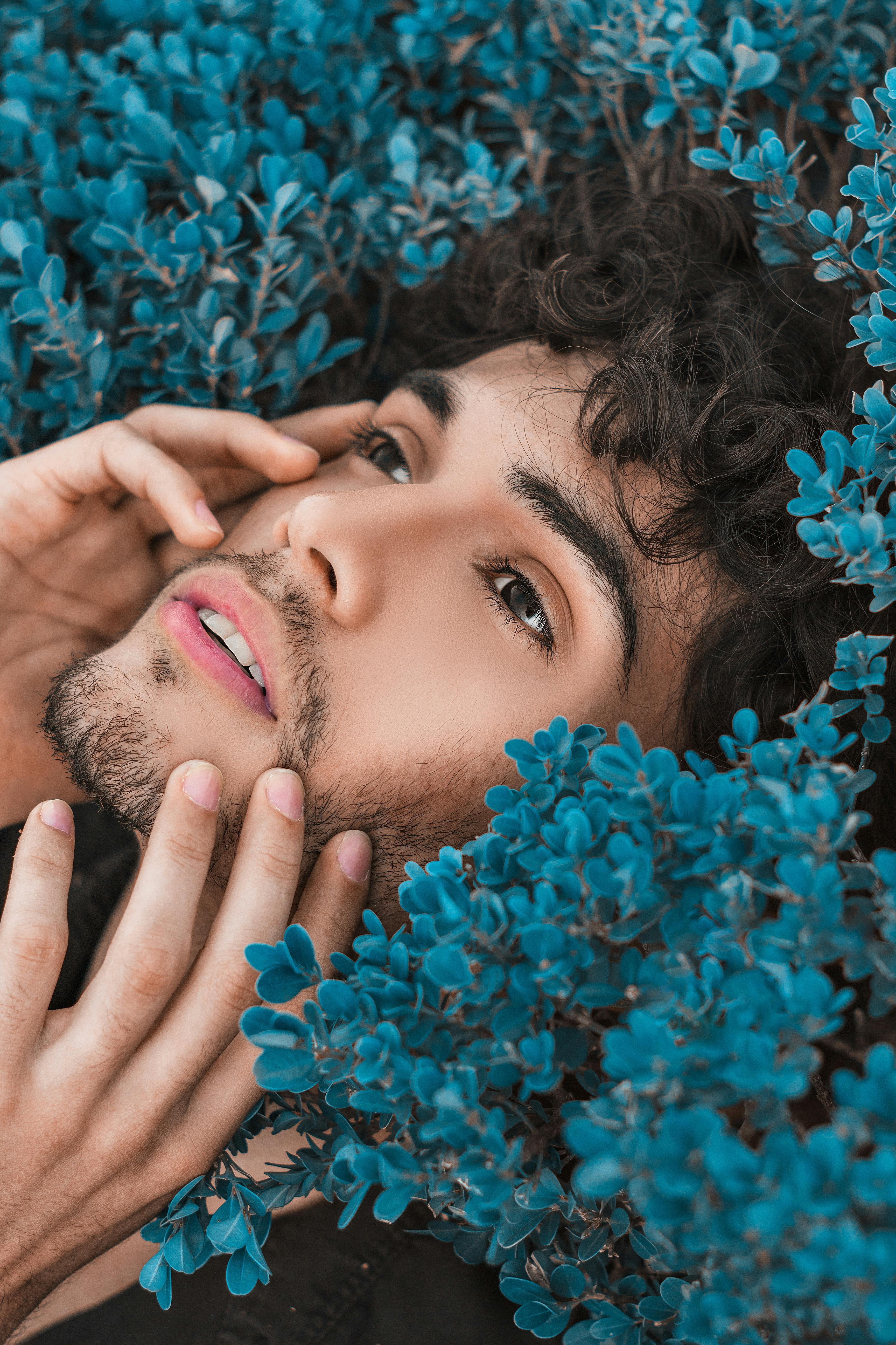 A person with curly dark hair and a trimmed beard lies surrounded by bright blue leaves, gently touching their face with their hands. They have soft, natural makeup and a contemplative expression.