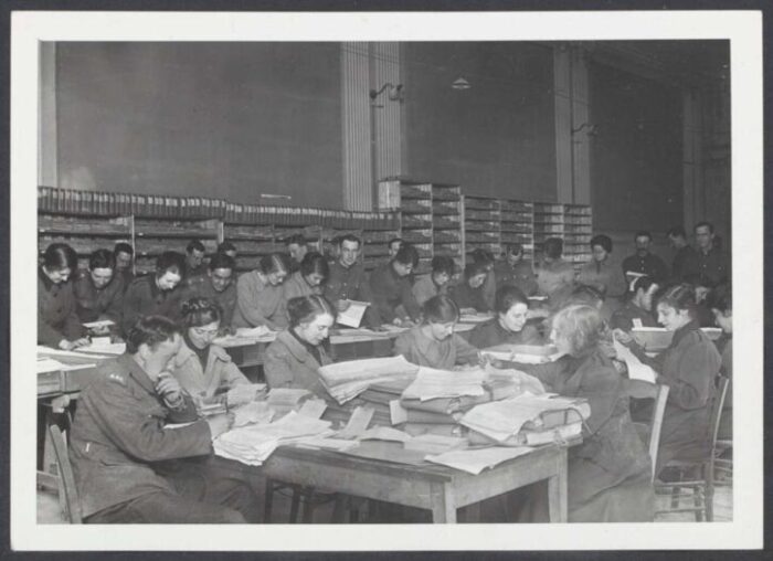 Dozens of Women’s Army Auxiliary Corps clerks seated at long tables, sorting and reviewing stacks of military documents in a large registry office during World War I.