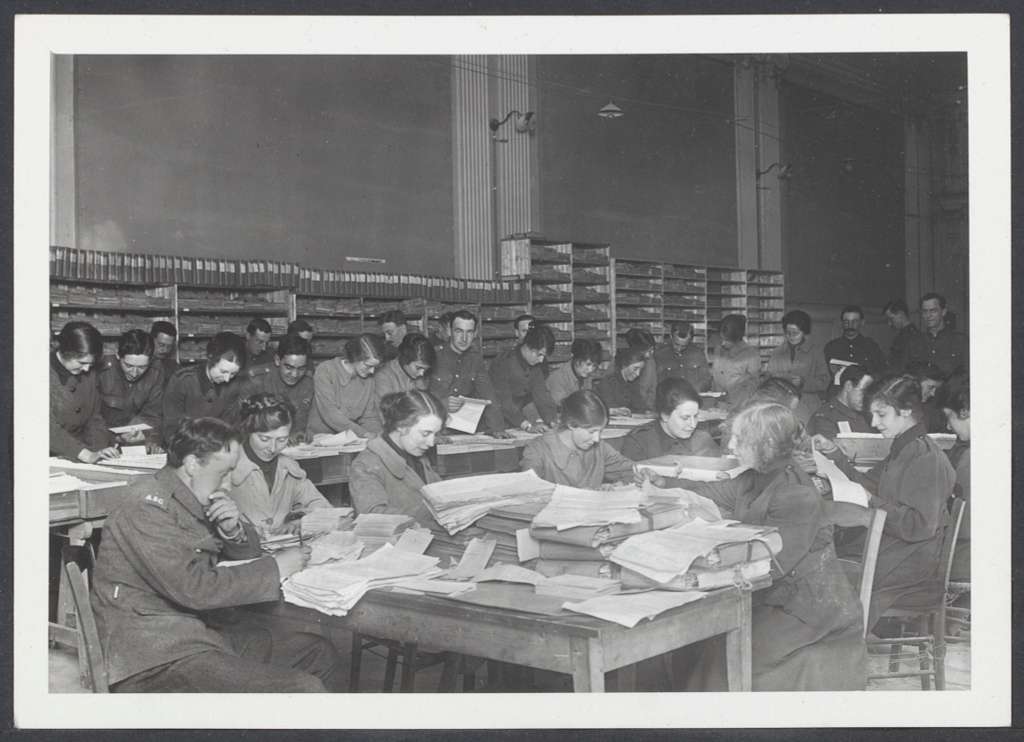Dozens of Women’s Army Auxiliary Corps clerks seated at long tables, sorting and reviewing stacks of military documents in a large registry office during World War I.