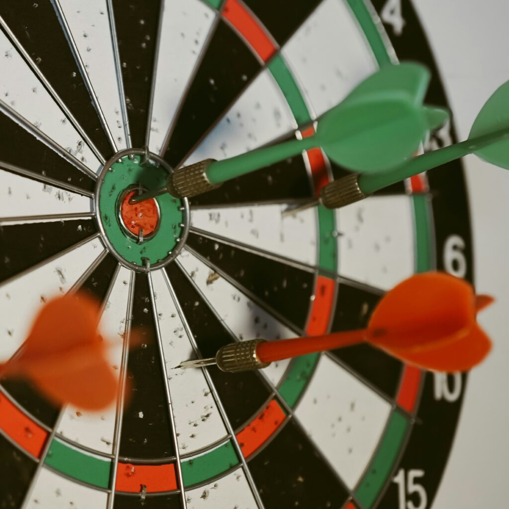 A close-up of a dartboard with several darts embedded near the bullseye. Two green darts and two orange darts are clustered tightly around the center, showing precise aim. The board’s black, white, red, and green sections are worn from frequent use, suggesting practice and focus.
