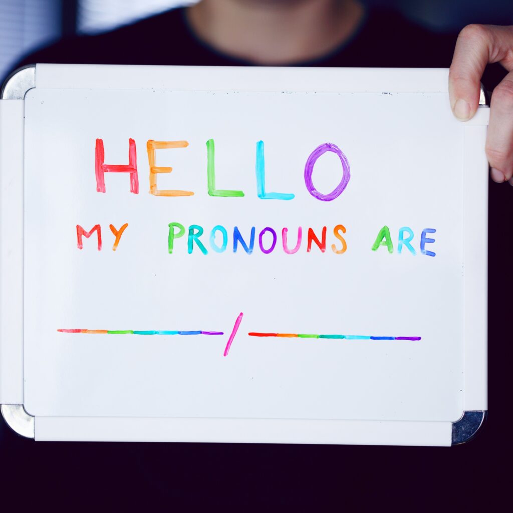 A person holds up a whiteboard with colorful rainbow text that reads “HELLO MY PRONOUNS ARE” followed by a blank line and a slash. The sign is written in vibrant marker colors representing inclusivity and pride, emphasizing the importance of sharing and respecting personal pronouns.