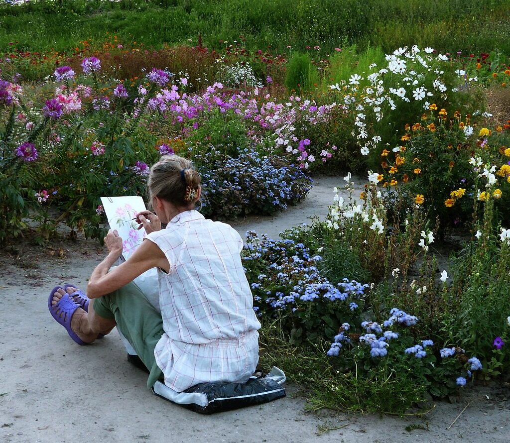 A young woman artist stands at an easel, painting on a canvas in a sunlit studio. She wears a casual shirt and jeans, with her hair tied back, focusing on her artwork. The room is filled with natural light, art supplies, and soft neutral tones, creating a calm and creative atmosphere.
