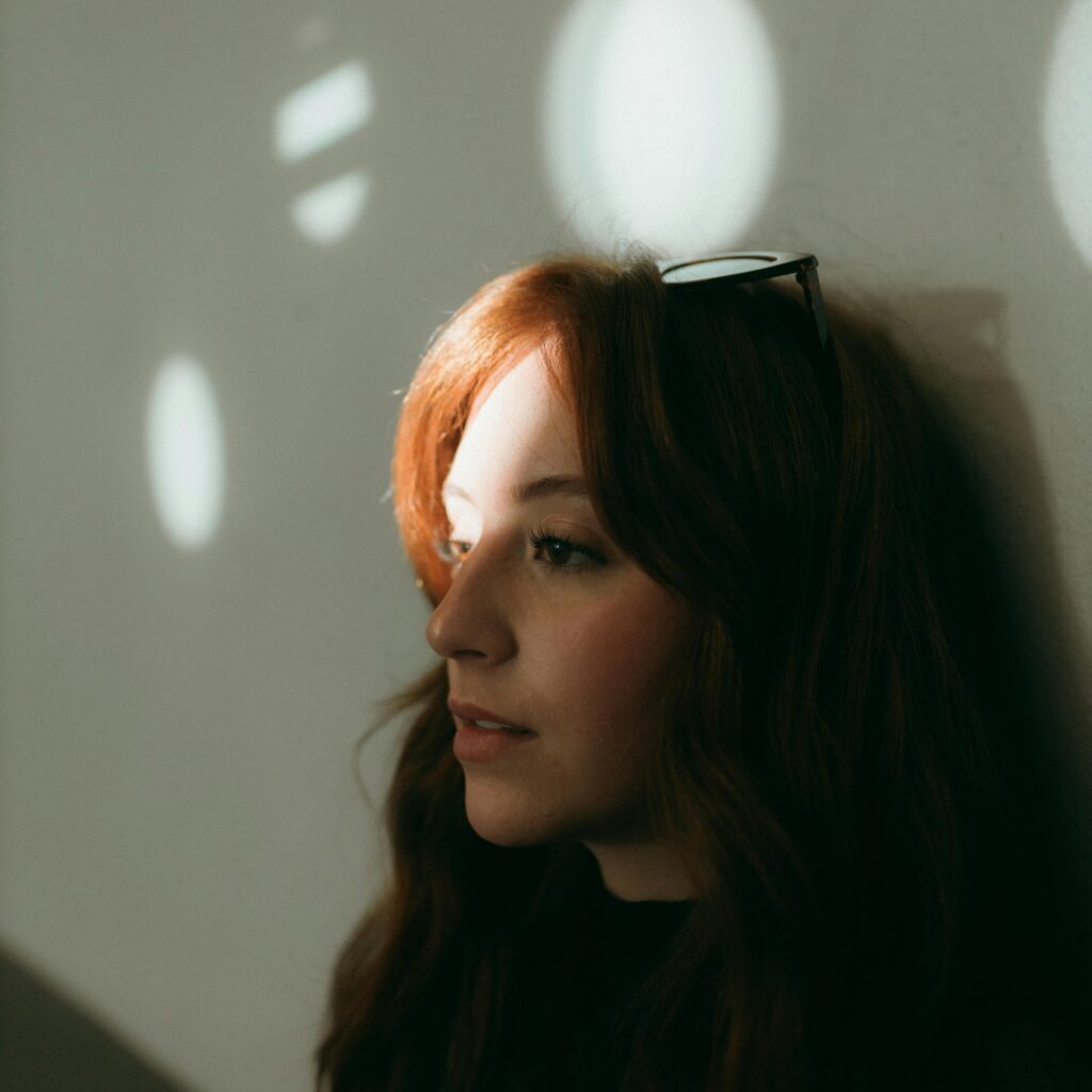 A young woman with long reddish-brown hair leans against a wall, her face softly lit by patches of sunlight filtering through a nearby window. She wears dark clothing and a pair of sunglasses rests on top of her head. The mood is calm and introspective, with warm light and gentle shadows creating a serene, contemplative atmosphere.