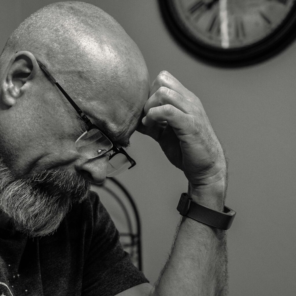 A middle-aged man with glasses and a beard sits with his head bowed, resting his forehead against his hand in a thoughtful or stressed pose. The black-and-white image highlights the texture of his skin and the intensity of the moment. A clock on the wall behind him suggests the passage of time, adding to the reflective mood.