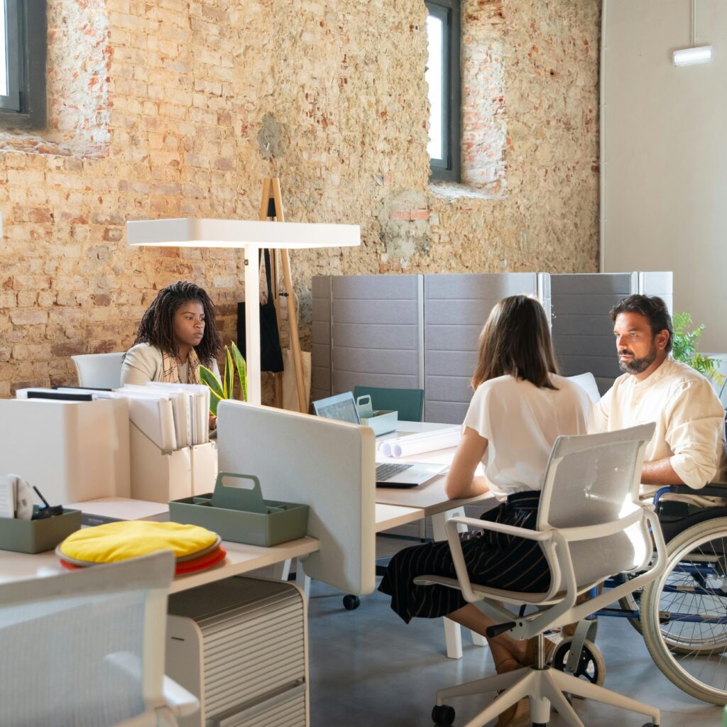 A group of students sits together at a large wooden table in a bright, modern study area. They are using laptops, notebooks, and tablets while discussing ideas. Shelves with books and plants line the background, and large windows let in natural light, creating a warm, collaborative atmosphere.