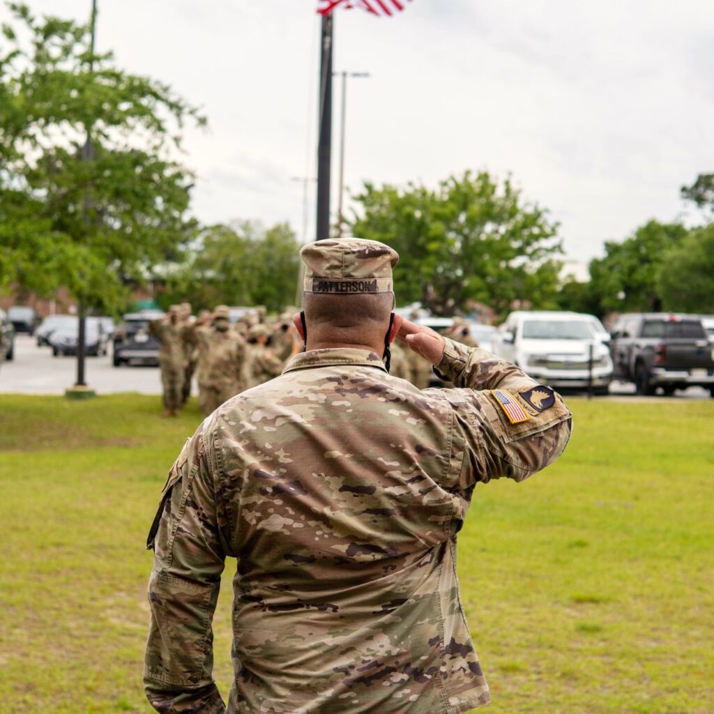Soldier saluting an American flag at half-staff on a grassy field with other service members in the background