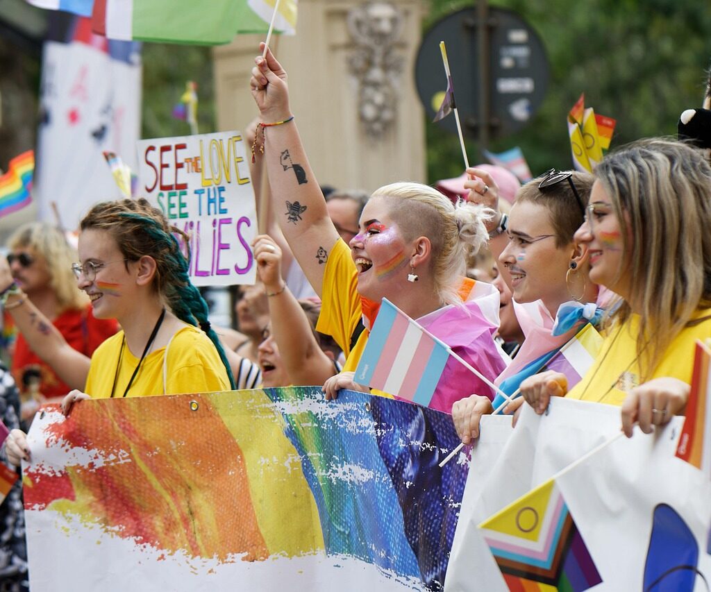 A group of smiling marchers at a Pride parade holding a rainbow banner, transgender and Progress Pride flags, and a sign reading "See the love, see the families."