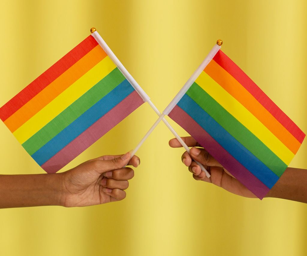 Two hands holding rainbow Pride flags crossed together against a shimmery gold backdrop.